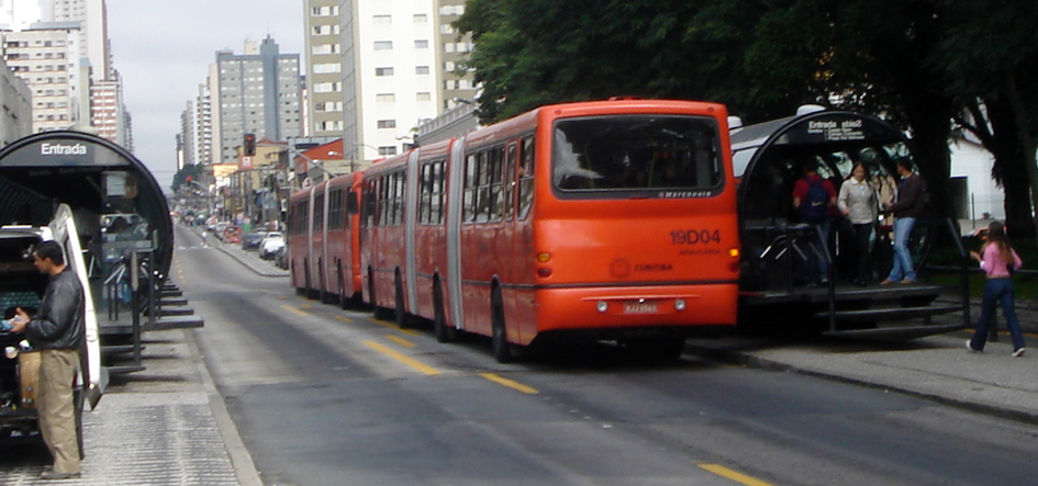 Red Marcopolo Torino bi-articulated buses on Volvo B10M chassis boarding passengers at a raised tube-shaped station on Curitiba's RIT bus rapid transit network.
