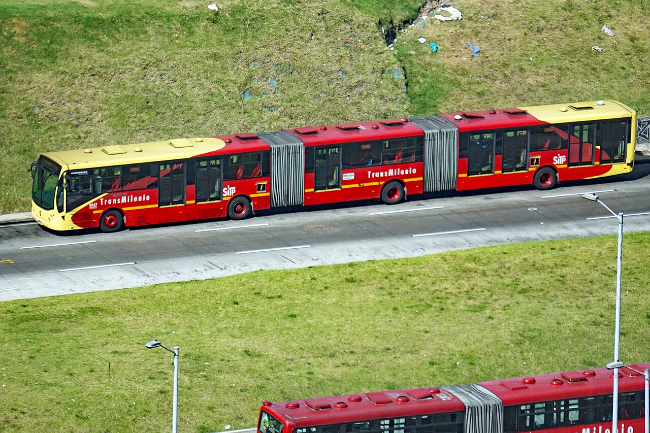 A red bi-articulated TransMilenio bus on a dedicated busway in Bogotá, Colombia, stretching across three sections.